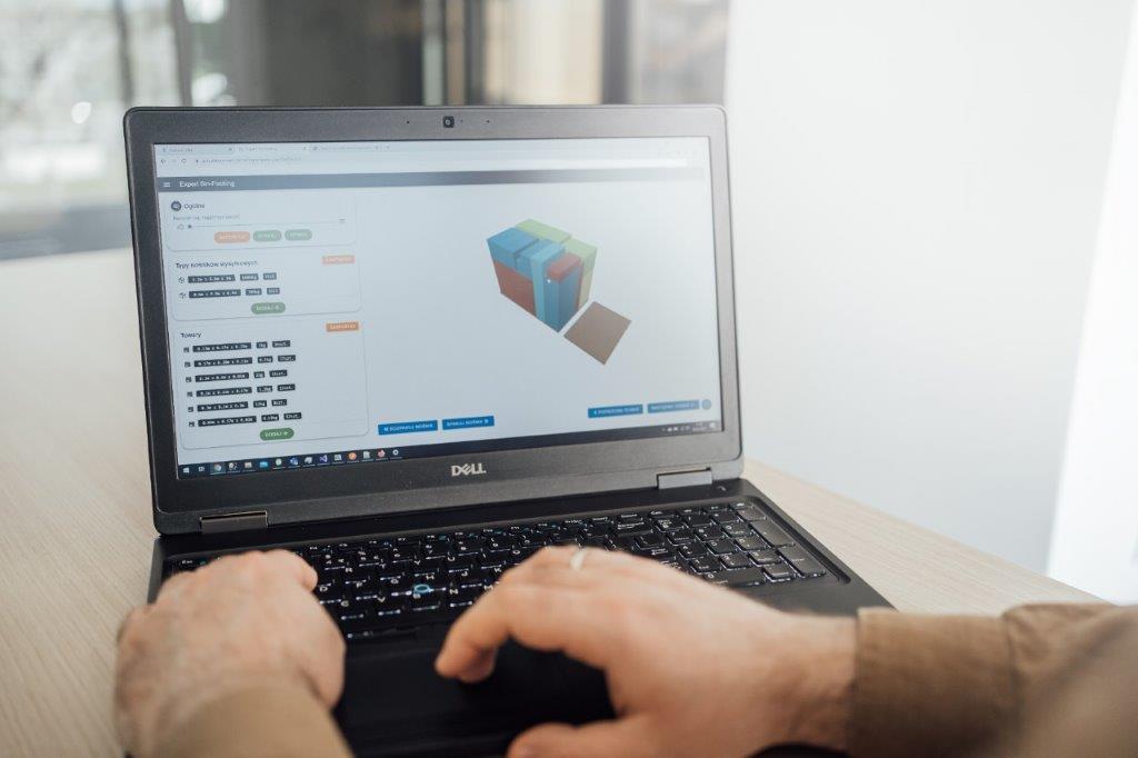 Na zdjęciu widać osobę pracującą przy laptopie, na którego ekranie wyświetlany jest interfejs systemu do zarządzania magazynem. W tle znajdują się elementy przestrzeni biurowej. Fotografia przedstawia wykorzystanie nowoczesnego oprogramowania wspierającego organizację i optymalizację procesów magazynowych.