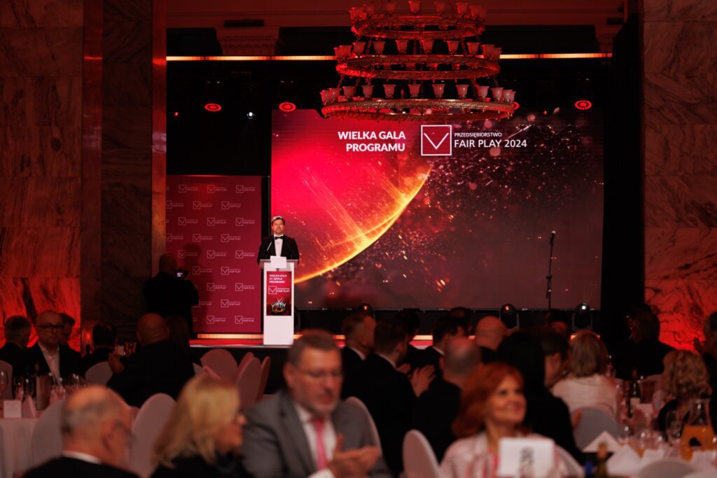 Na zdjęciu widać Wielką Galę Programu „Przedsiębiorstwo Fair Play 2024”. Na scenie znajduje się prowadzący wydarzenie, a w tle wyświetlana jest oprawa graficzna programu. W eleganckiej sali zgromadzili się przedstawiciele firm i zaproszeni goście, uczestniczący w uroczystej gali przy stołach bankietowych. Całość utrzymana jest w odświętnej, czerwono-czarnej oprawie.