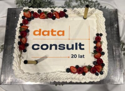 Na zdjęciu przedstawiono prostokątny tort jubileuszowy przygotowany z okazji 20-lecia firmy Data Consult. Tort ozdobiono białym kremem, świeżymi owocami – truskawkami, malinami i borówkami – oraz dekoracyjnymi fontannami tortowymi. Na środku znajduje się logo firmy oraz napis „20 lat”, podkreślający rocznicę działalności przedsiębiorstwa.
